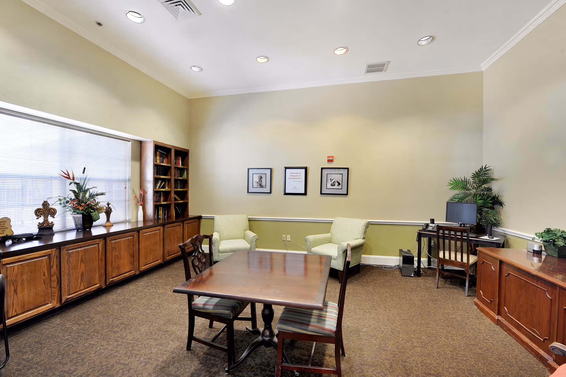 A well-lit room with beige walls and carpeted floor featuring a wooden table with four chairs in the center. Against the left wall is a long wooden cabinet with decorative items and a bookshelf filled with books. Two light green armchairs are placed against the far wall, which has three framed pictures. On the right side, there is a wooden desk with a computer and a chair, along with some potted plants.