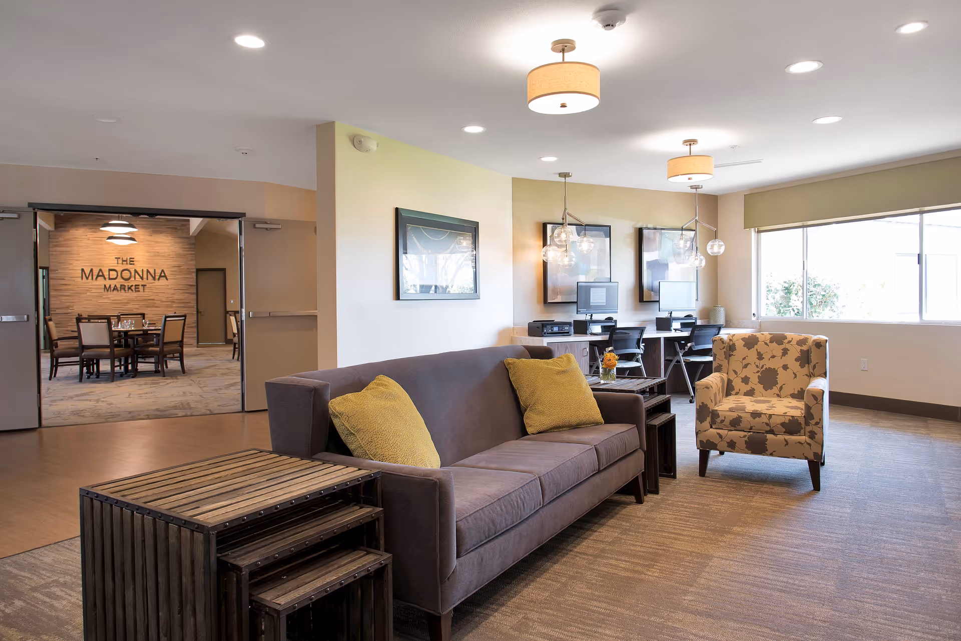 A well-lit common area with a gray sofa adorned with two yellow pillows, a floral-patterned armchair, and a set of nested wooden tables. In the background, there are two desks with computers and office chairs, framed artwork on the walls, and large windows letting in natural light. To the left, double doors open to a dining area labeled 'The Madonna Market' with a round dining table and chairs.