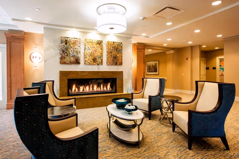 A cozy seating area in a senior living facility with four high-back armchairs arranged around two oval coffee tables. The room features a modern gas fireplace set in a stone surround, with three abstract art pieces hanging above it. The walls are painted in warm neutral tones, and the space is softly lit by ceiling lights and wall sconces.