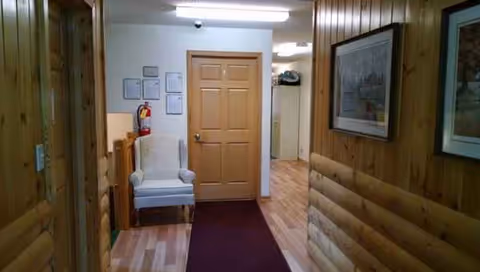 Wood-paneled hallway with a chair, framed pictures, door, and burgundy runner rug.