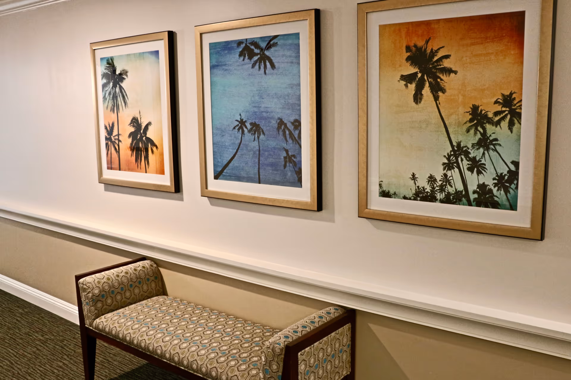 A hallway wall decorated with three framed pictures of palm trees against colorful skies in shades of orange, blue, and green. Below the pictures is a cushioned bench with patterned upholstery and wooden armrests.