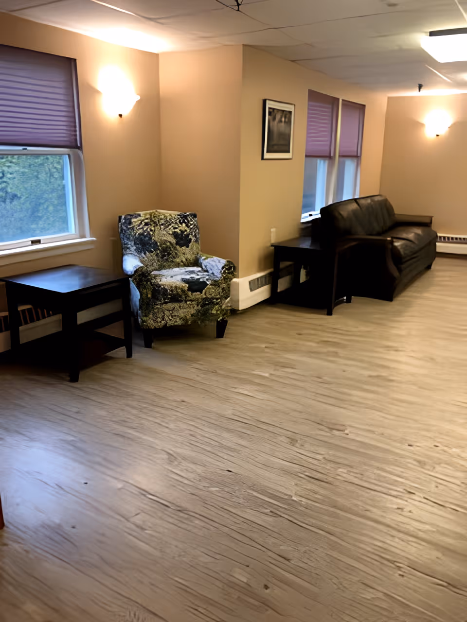 A living room area with beige walls and wood-patterned flooring. There is a patterned armchair and a dark wooden side table near a window with purple blinds on the left. On the right side, there is a dark leather sofa with another dark wooden side table next to it. The room is softly lit by wall-mounted lights.