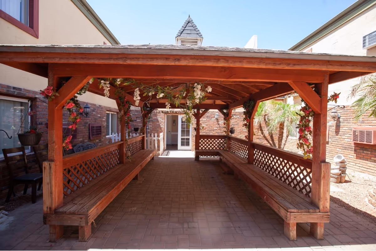 Covered wooden gazebo with benches on both sides, decorated with hanging flowers, located in an outdoor courtyard area with brick walls and some plants visible.