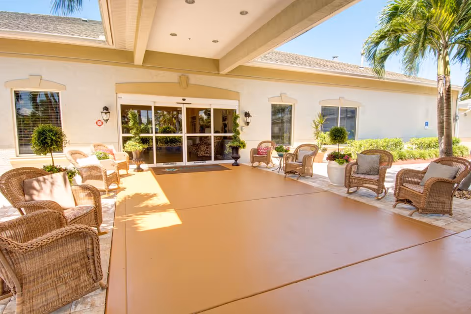 Covered entrance patio with wicker chairs and potted plants leading to glass automatic doors of a senior living facility.