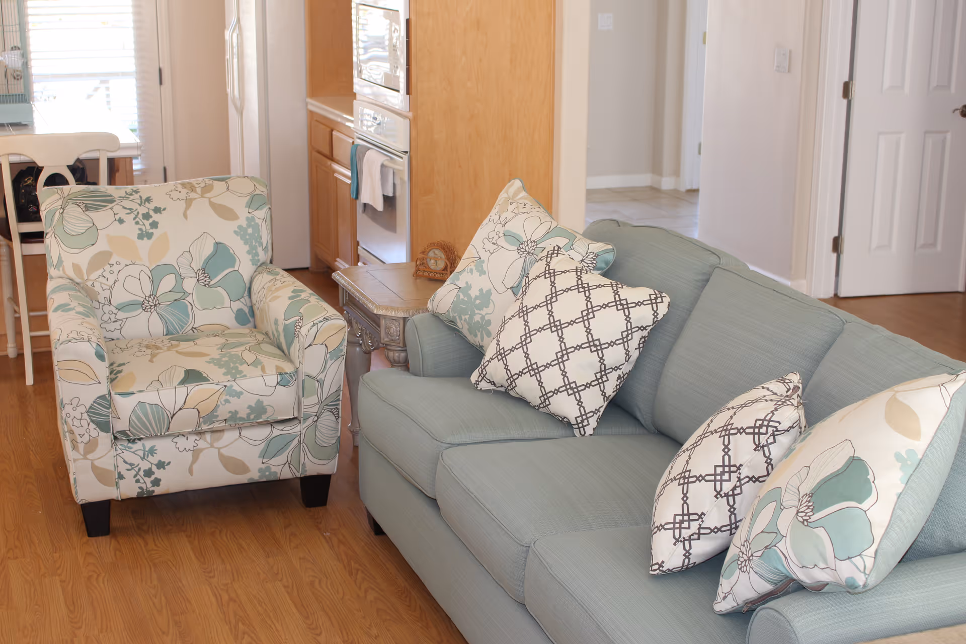 A cozy living room area featuring a light blue sofa with patterned throw pillows and a floral armchair. The room has wooden flooring and an adjacent kitchen area with wooden cabinets and an oven. A small side table is placed between the sofa and armchair.