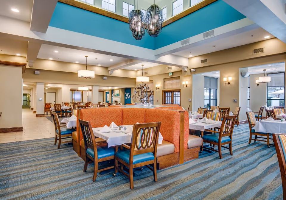 A spacious dining room with multiple tables covered in white tablecloths and set with napkins and silverware. The room features a large orange booth seating area in the center, wooden chairs with blue cushions, and modern light fixtures hanging from the ceiling. The walls are painted beige with blue accents near the ceiling, and large windows allow natural light to enter.