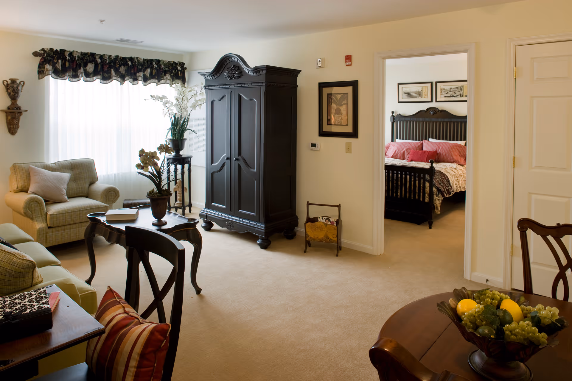 A cozy living room with beige carpet and cream-colored walls, featuring a plaid armchair with a pillow, a dark wooden armoire, a small table with a potted plant, and a wooden dining table with a bowl of fruit. Through an open doorway, a bedroom with a dark wooden bed frame, red pillows, and framed pictures on the wall is visible.