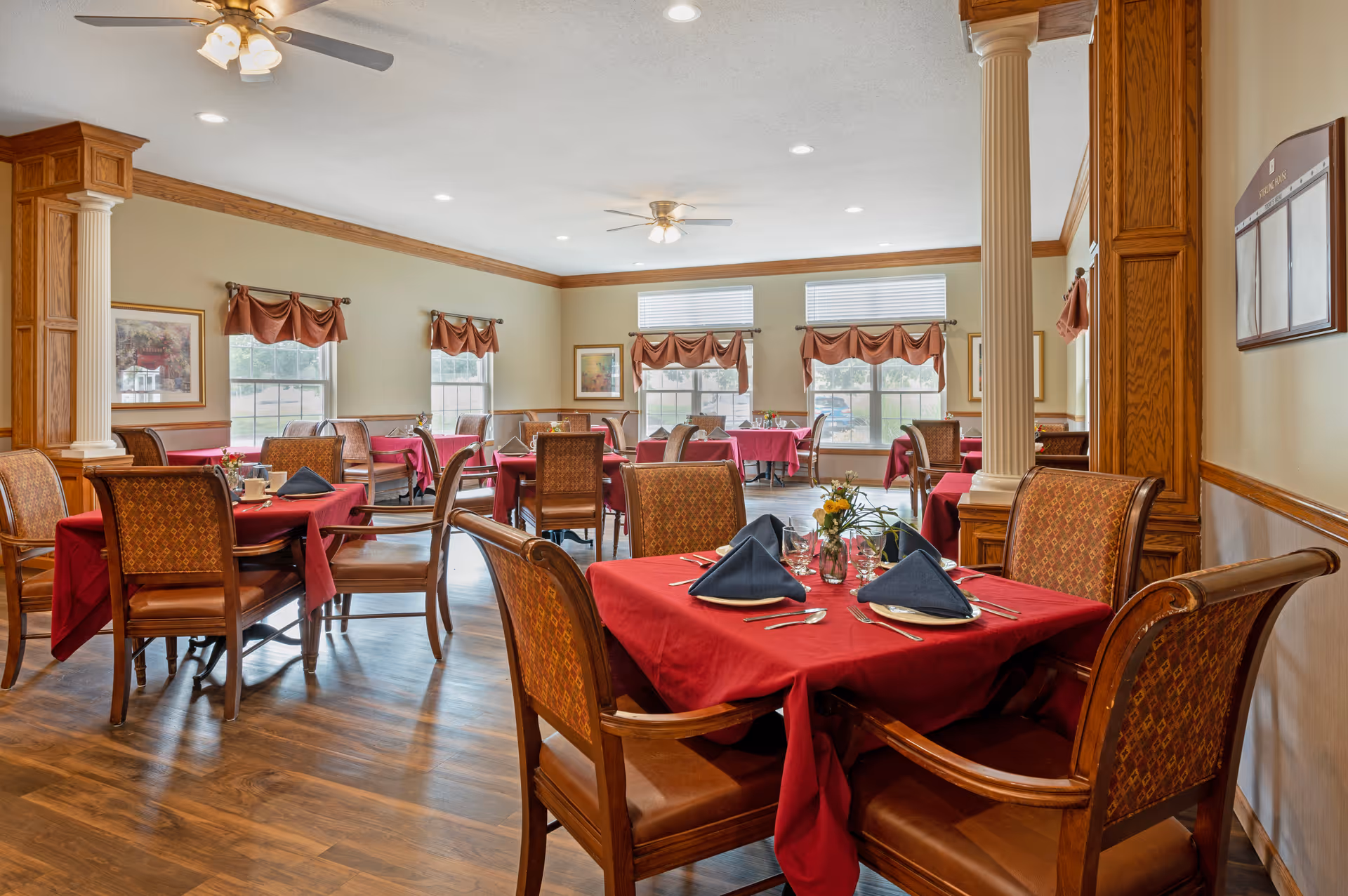 A bright dining room with multiple tables covered in red tablecloths and set with plates, silverware, glasses, and folded navy blue napkins. The room features wooden chairs with patterned upholstery, large windows with brown valances, ceiling fans with lights, and wooden columns and trim.