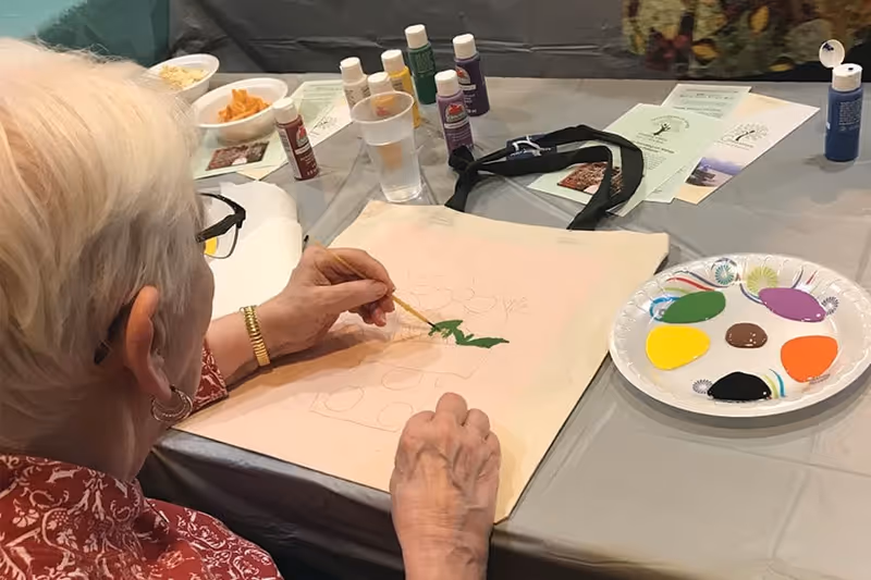 An elderly person with short white hair and glasses is painting on a canvas tote bag at a table covered with a gray tablecloth. Various colors of paint are on a paper plate nearby, along with several bottles of paint, a cup of water, and some papers on the table.
