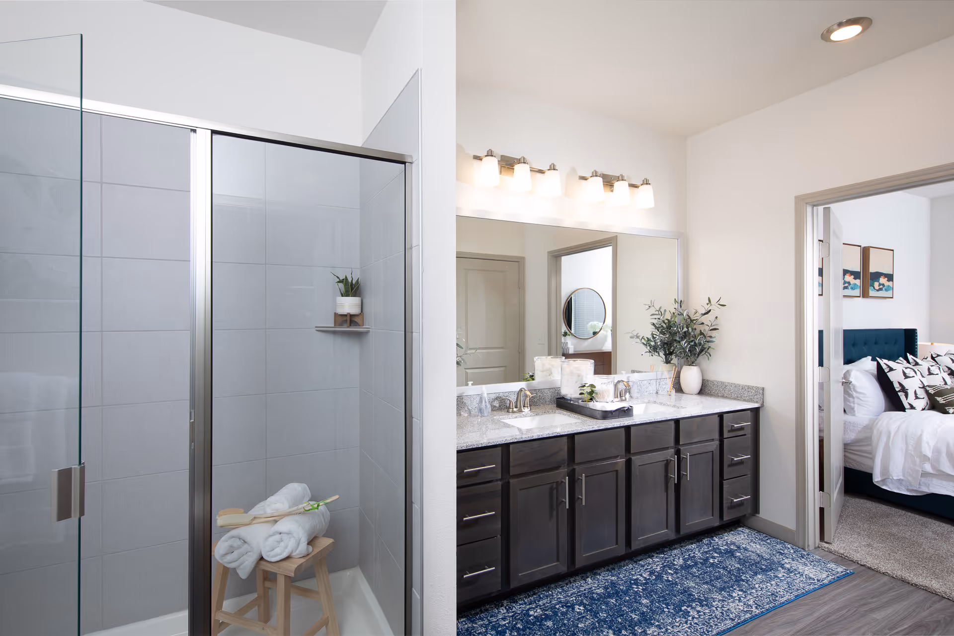 Modern bathroom with a glass-enclosed shower featuring gray tiles and a wooden stool with rolled white towels. A double sink vanity with dark wood cabinets, granite countertop, and a large mirror with six light fixtures above. A blue patterned rug is on the floor. The bathroom opens into a bedroom with a bed, pillows, and wall art visible.