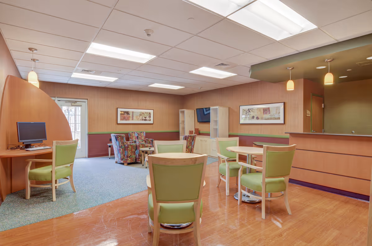A senior living facility common area with light wood flooring and green cushioned chairs around small round tables. There is a computer workstation with a green chair on the left side, and a seating area with colorful patterned armchairs and a TV mounted on the wall in the background. The walls are decorated with framed artwork, and pendant lights hang from the ceiling. A reception desk is visible on the right side.