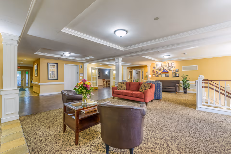 A spacious and well-lit common area in a senior living facility with beige walls and carpeted floors. The room features a red sofa with patterned cushions, two dark brown leather chairs, a wooden coffee table with a vase of colorful flowers, and a gray sofa in the background. There are framed pictures on the walls, a potted plant, and white pillars supporting the ceiling. A staircase with white railings is visible on the right side, and an elevator and hallway are seen in the background.