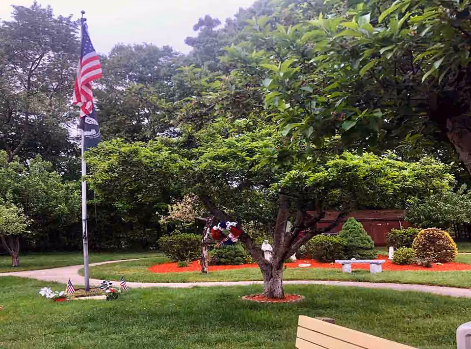 A peaceful outdoor garden area with a flagpole displaying the American flag and another flag beneath it. The garden features green grass, a variety of trees and bushes, a stone bench, and a winding paved pathway. There are red mulch beds around the trees and bushes, and a wooden shed is visible in the background.