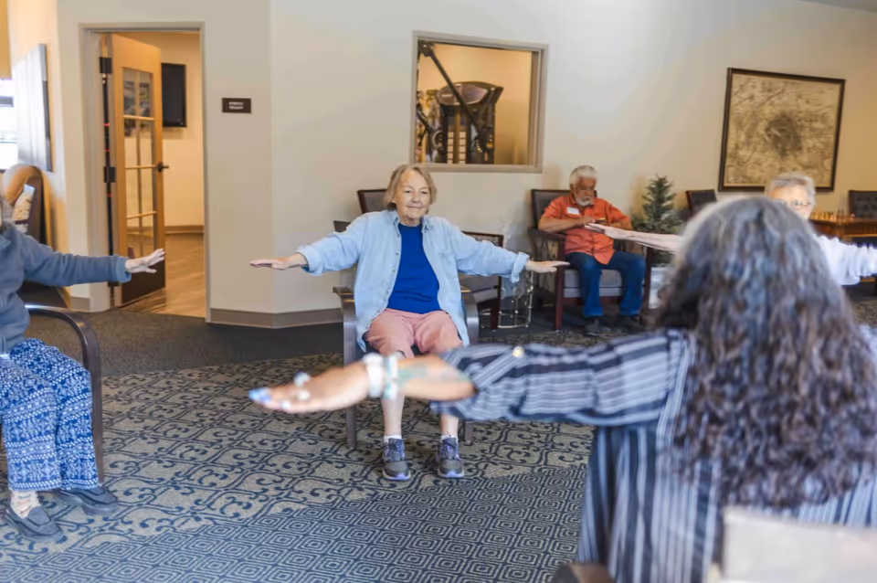 A group of elderly individuals seated in chairs in a common room, participating in a seated exercise session with arms extended to the sides. The room has patterned carpet, light-colored walls, and some framed artwork and furniture in the background.