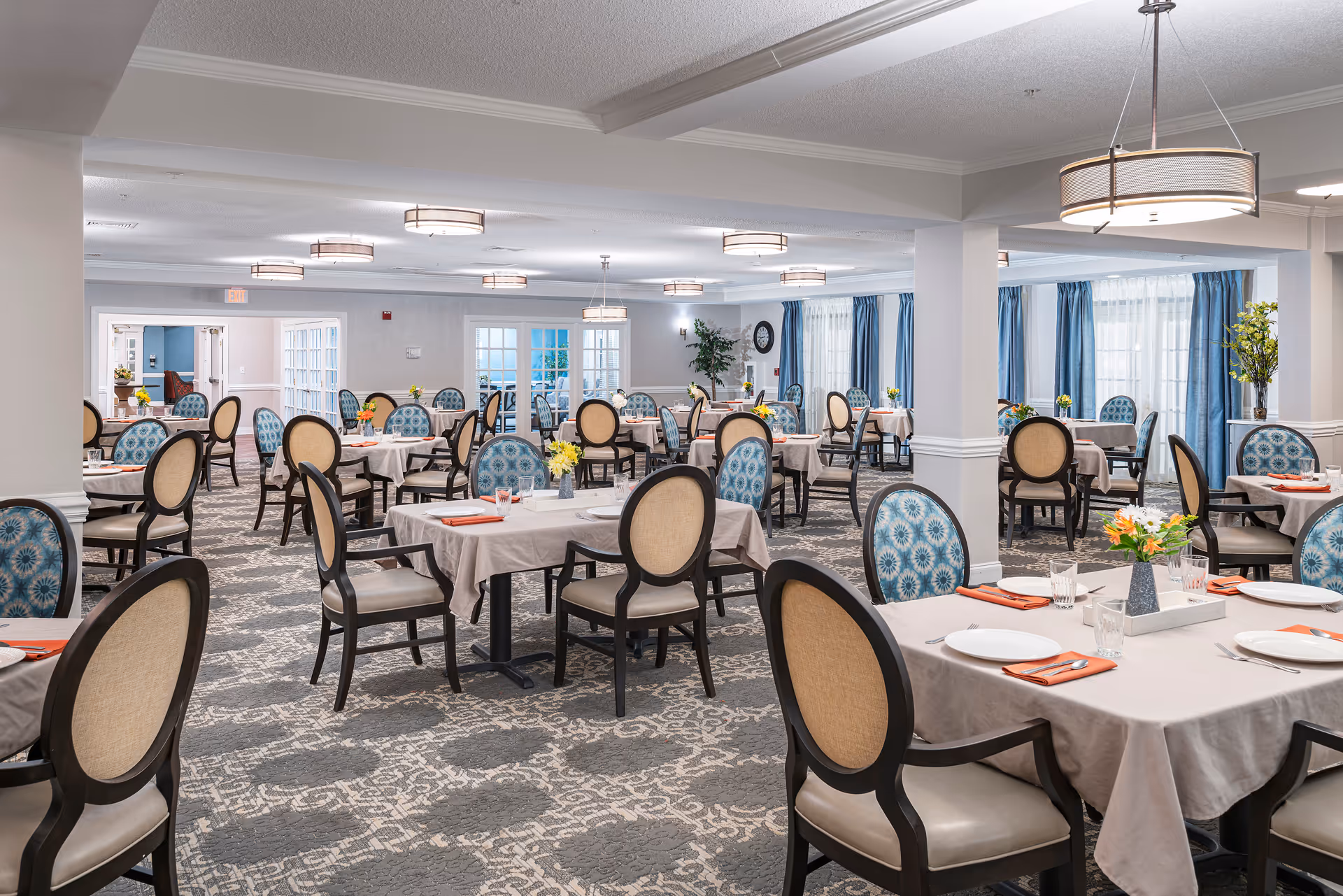 A spacious dining room with multiple tables covered in beige tablecloths, each set with plates, glasses, and orange napkins. The chairs have dark wooden frames with beige and blue patterned upholstery. The room has large windows with blue curtains, modern ceiling lights, and a patterned carpet floor.