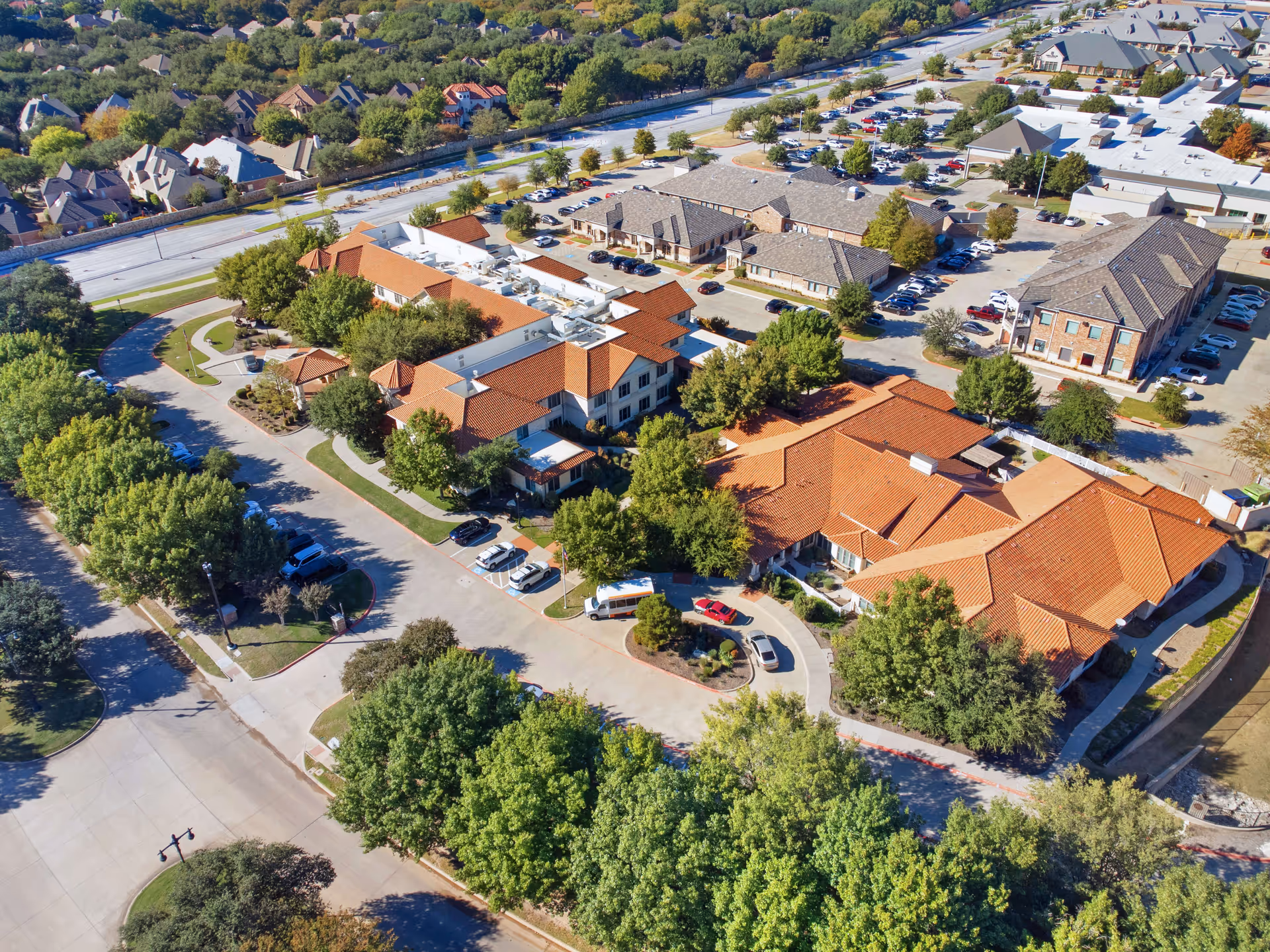 Aerial view of Sunrise of Frisco senior living facility showing multiple buildings with red-tiled roofs, surrounded by trees, parking lots with cars, and nearby residential neighborhood.