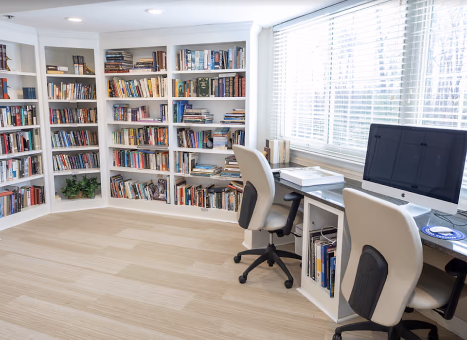 A bright room with large windows covered by white blinds, featuring white bookshelves filled with books along the walls. In front of the windows, there is a long desk with two white office chairs on wheels and an iMac computer on the desk.