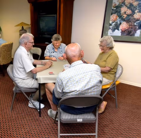Four elderly people sitting around a table playing cards in a room with carpeted floor, a wooden cabinet with a TV, a lamp, and a framed floral painting on the wall.