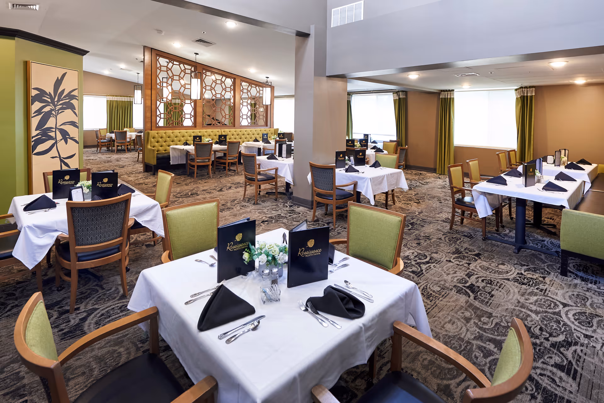 A spacious dining room with multiple tables covered in white tablecloths, each set with black napkins, silverware, and menus labeled Renaissance. The room features green cushioned chairs, patterned carpet, large windows with green curtains, and decorative wooden partitions.