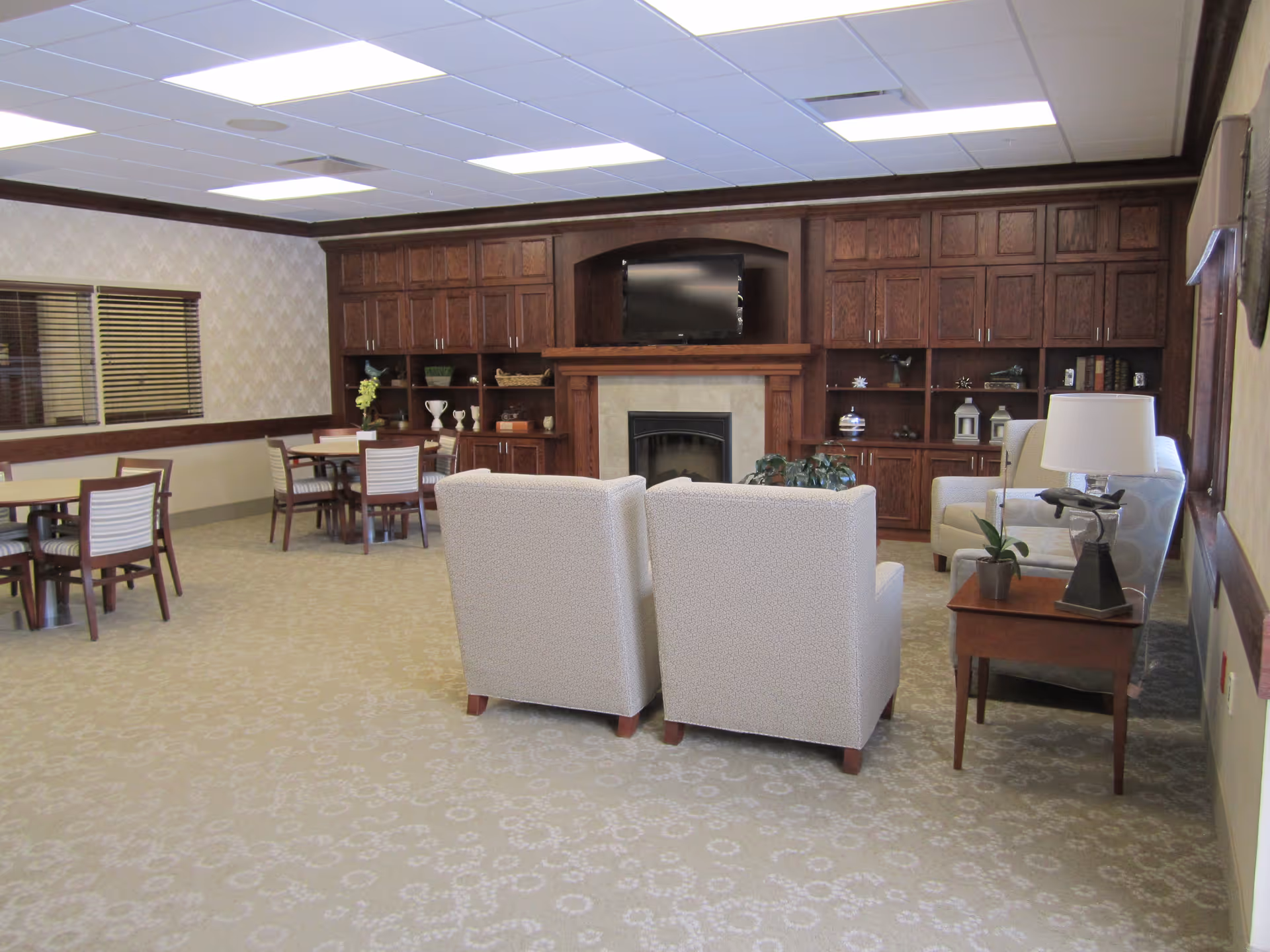 A cozy common area in a nursing and rehab center featuring two beige armchairs facing a fireplace with a mounted flat-screen TV above it. Surrounding the fireplace is a large wooden built-in cabinet with shelves displaying decorative items. To the left, there are several tables and chairs arranged for dining or socializing. The room has a patterned carpet, beige walls with a wooden trim, and a window with blinds.
