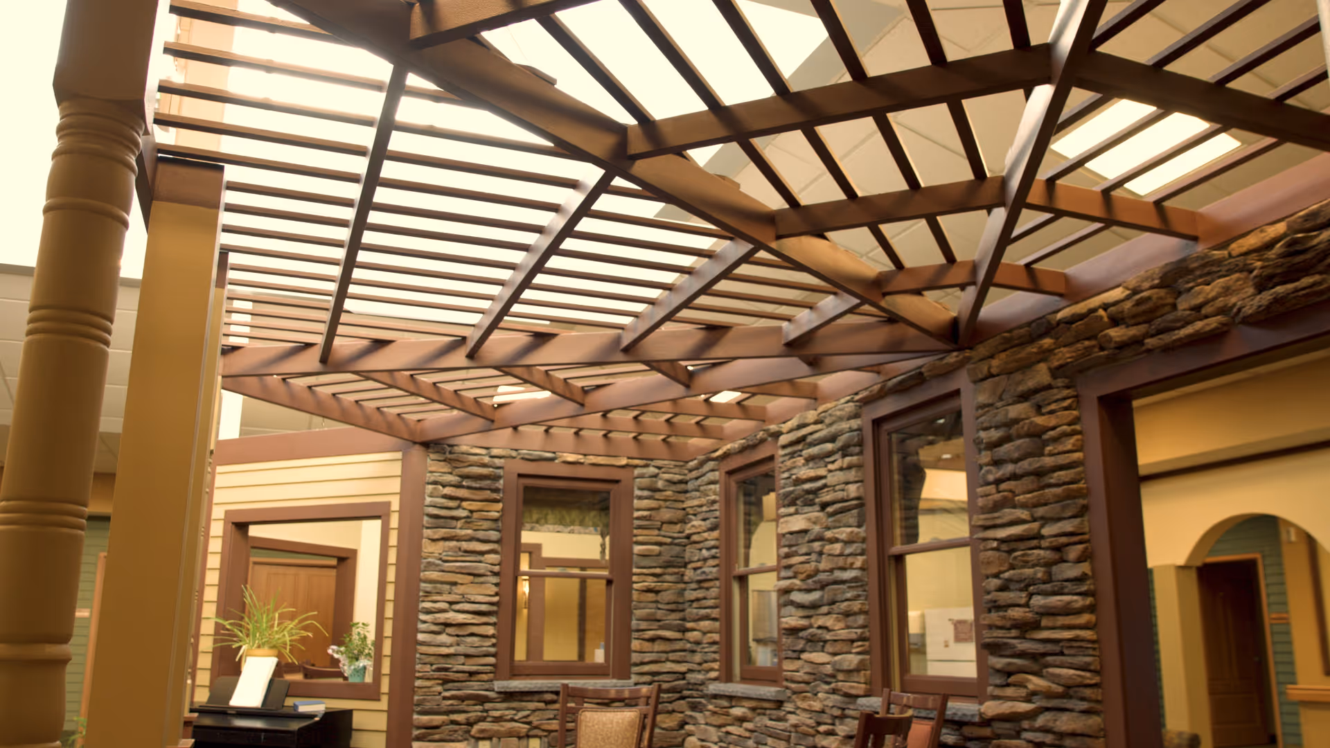 Indoor atrium with a wooden pergola ceiling, stone-clad walls, windows and chairs.