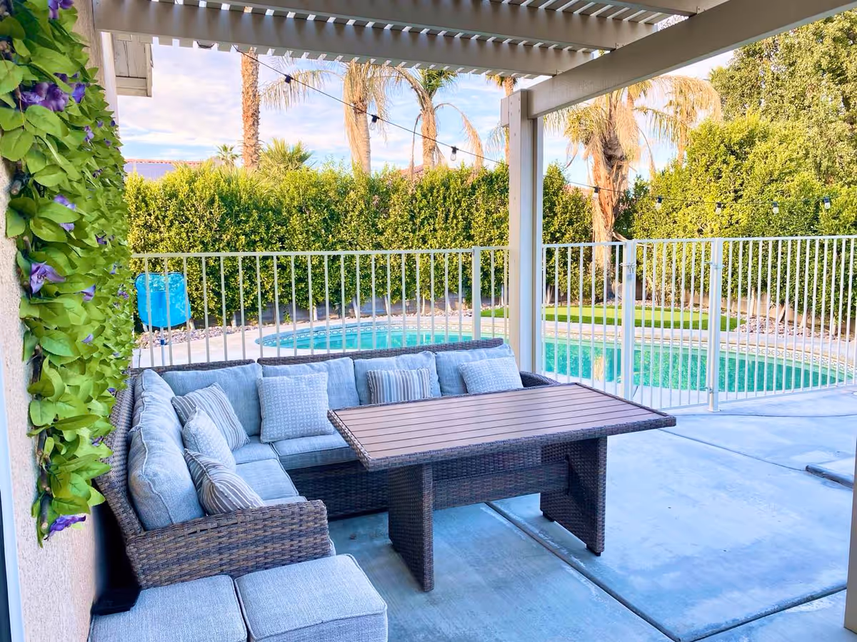 Outdoor patio area with a wicker sectional sofa with light blue cushions and a rectangular wooden table. The patio is covered with a pergola and overlooks a fenced swimming pool surrounded by greenery and palm trees.