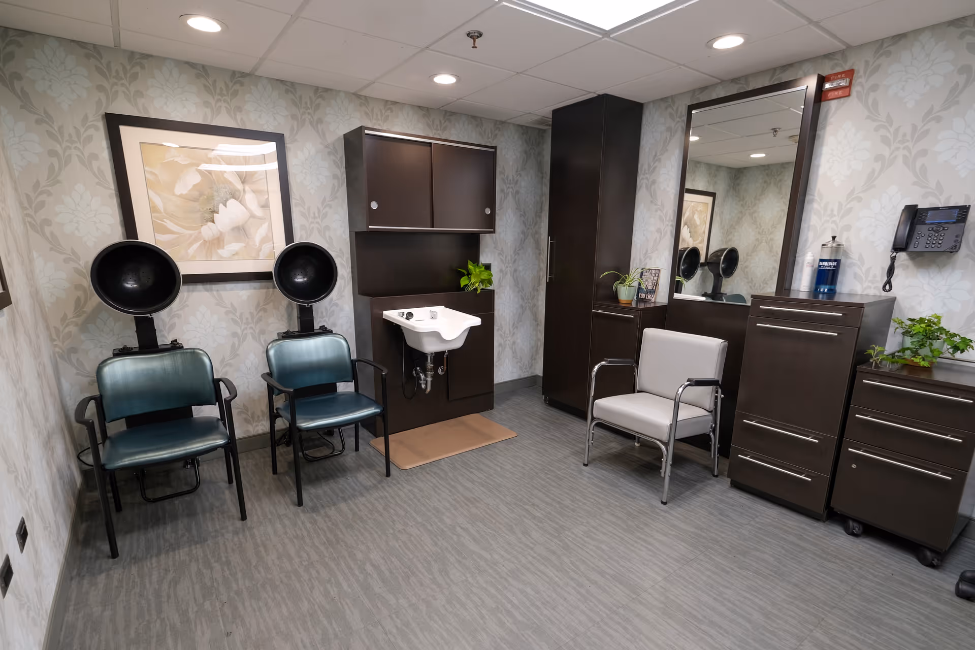 Interior salon room with two hooded hair dryers, a wash basin, chairs, a large mirror and dark cabinetry.