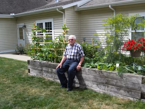 An elderly man wearing sunglasses and a plaid shirt sits on the edge of a raised wooden garden bed filled with various green plants and flowers. Behind him is a beige building with white-framed windows and a door. The scene is outdoors on a grassy lawn under daylight.