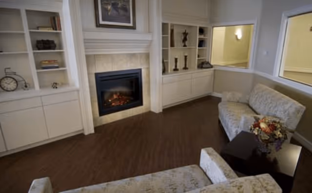 A cozy living room with two floral-patterned sofas facing each other, a dark wooden coffee table with a floral arrangement, built-in white shelves with decorative items, and a fireplace with a painting above it.