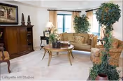 A cozy living room with a beige sofa, a patterned armchair, a round wooden coffee table with decorative items, a wooden cabinet, potted plants, and a window with curtains letting in natural light.
