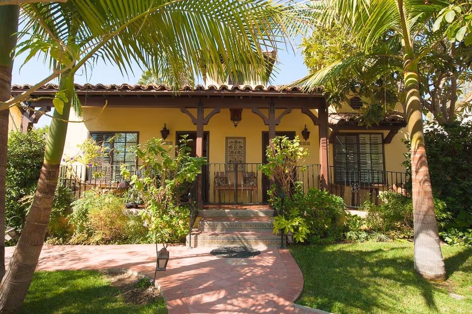 Front exterior of a yellow stucco house with a tiled roof, a small porch entrance, palm trees, and lush landscaping.
