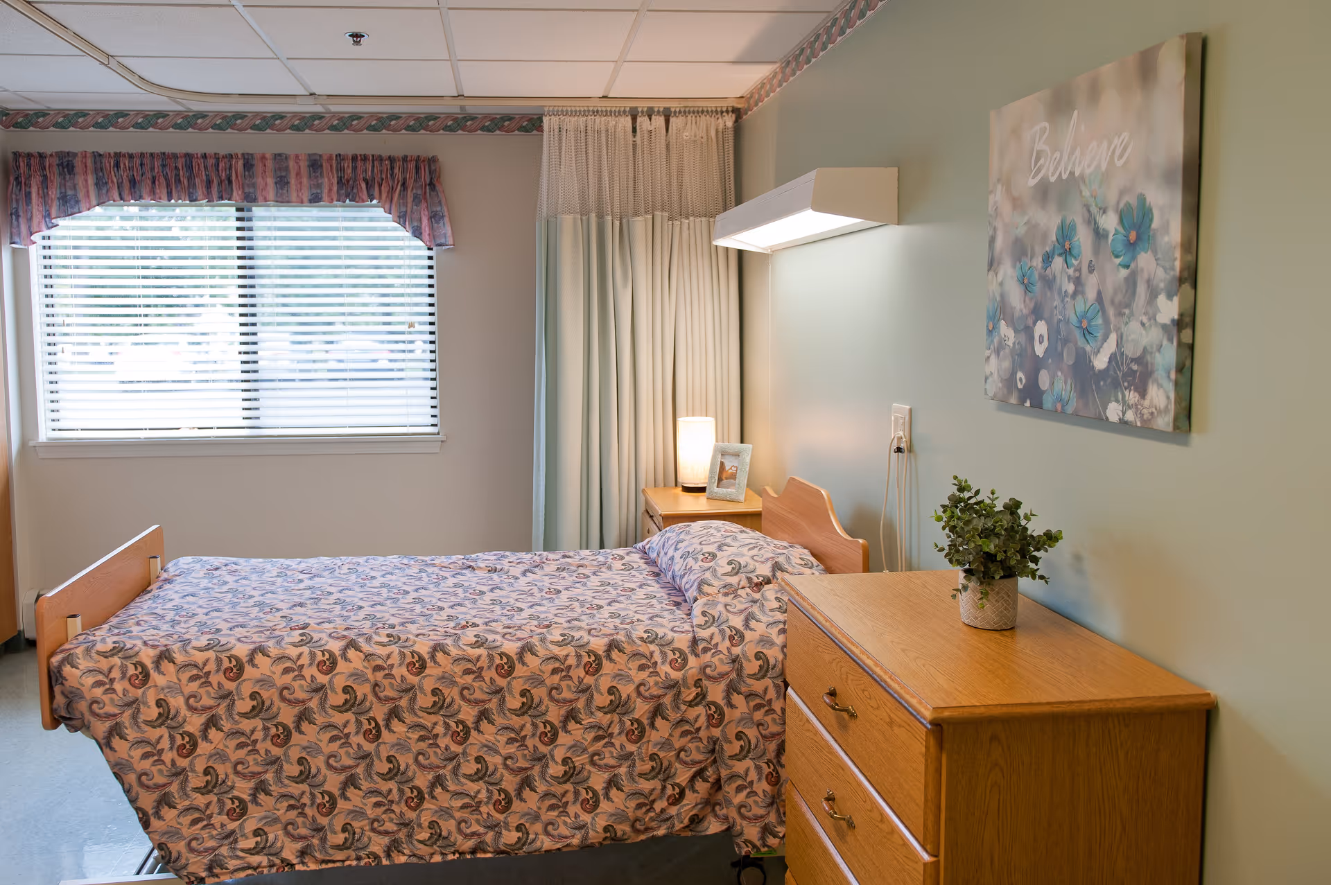 A single bed with a patterned bedspread in a senior living facility room. The room has a window with blinds and a floral valance, a wooden dresser with a potted plant on top, a nightstand with a lamp and a framed photo, and a wall-mounted light above the bed. A floral painting with the word 'Believe' hangs on the wall.