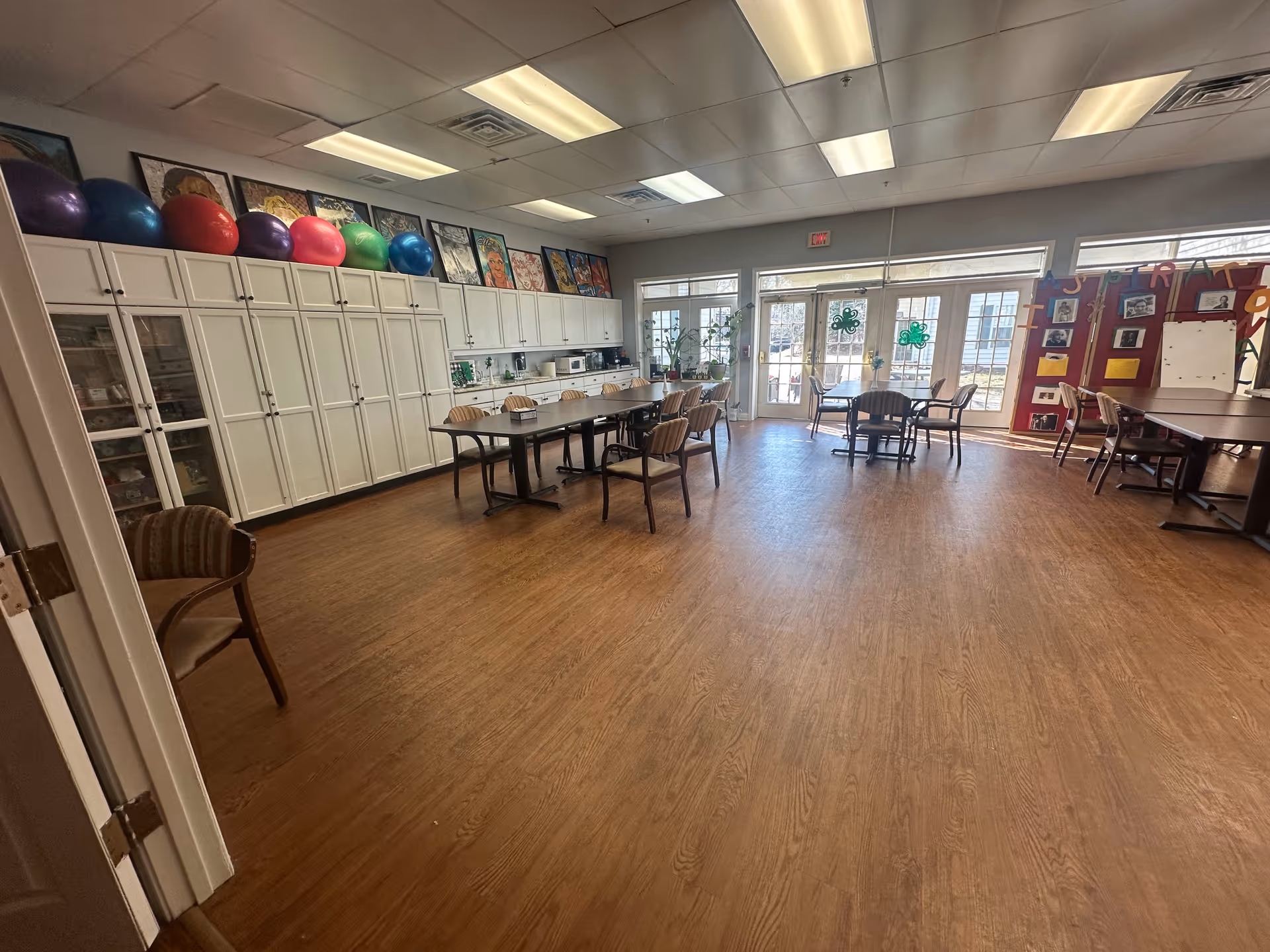 A spacious activity room with wooden flooring and white cabinets along one wall. Several colorful exercise balls are placed on top of the cabinets. There are multiple tables and chairs arranged around the room, with large windows and glass doors letting in natural light. The walls display various artworks and decorations.