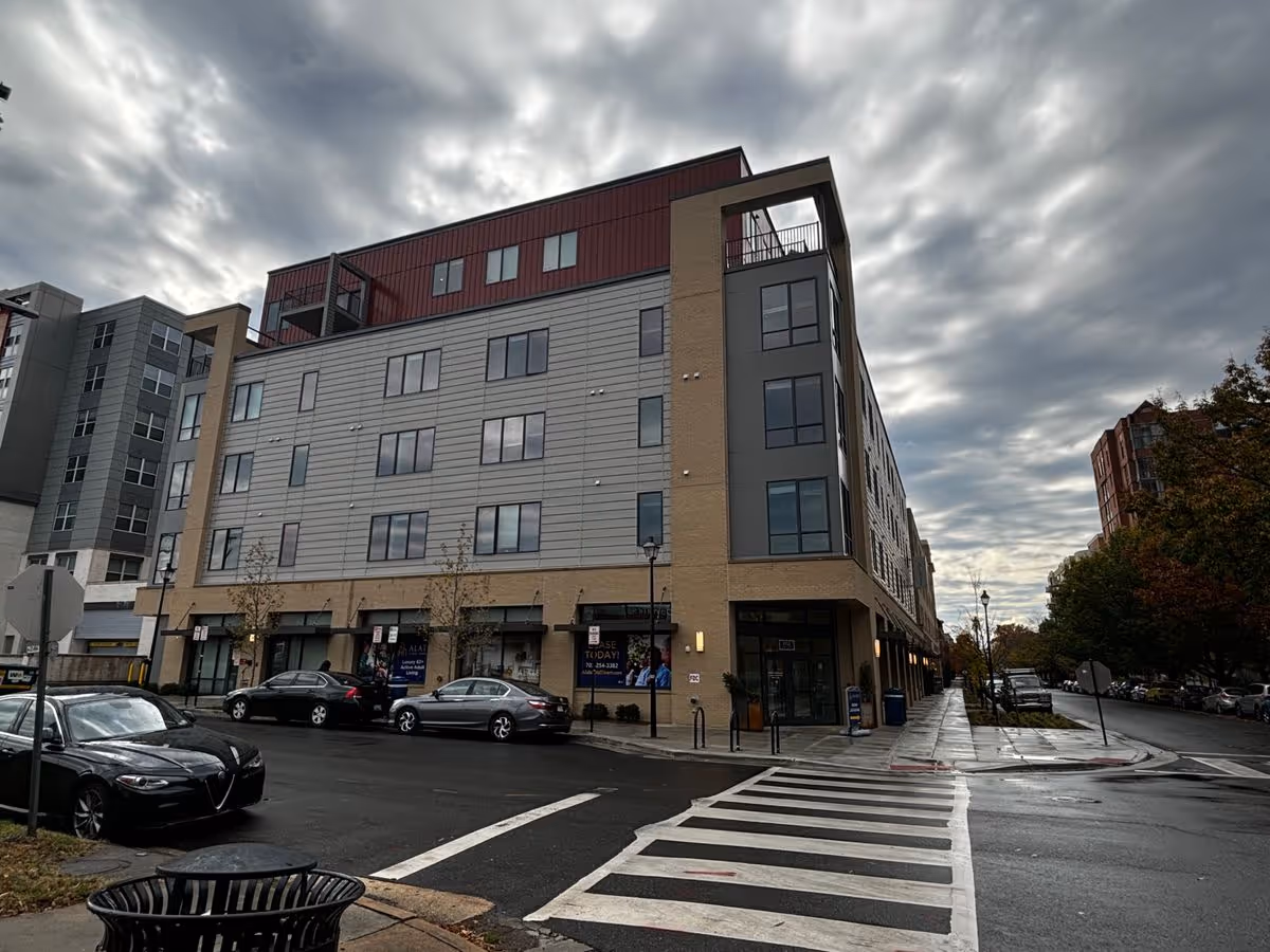 Corner view of a multi-story modern apartment building with ground-floor shops on a cloudy day.