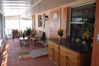 Covered outdoor patio area with cushioned chairs, a small table, potted plants, and a cabinet against the wall next to a door and window.