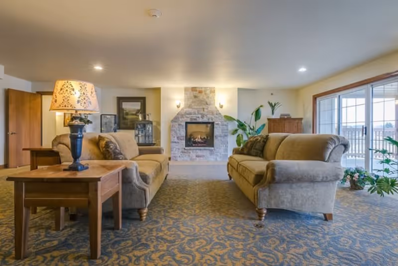 Spacious lounge with two sofas facing a central stone fireplace, side tables with a lamp, plants, and sliding glass doors.
