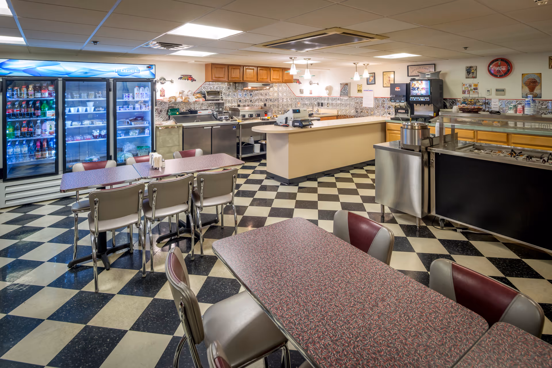 A retro-style dining area with checkered black and white flooring, several tables with maroon and gray chairs, a beverage cooler stocked with drinks, a counter with kitchen equipment, and a soda fountain machine in the background.