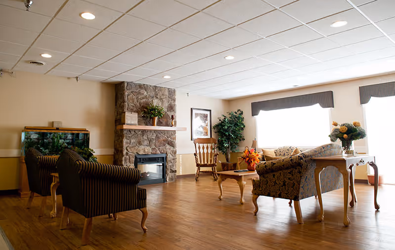 A cozy living room area with wooden flooring, a stone fireplace, two striped armchairs, a floral patterned sofa, a wooden rocking chair, a coffee table with a flower arrangement, and a side table with a vase of flowers. Large windows with valances allow natural light to fill the room.