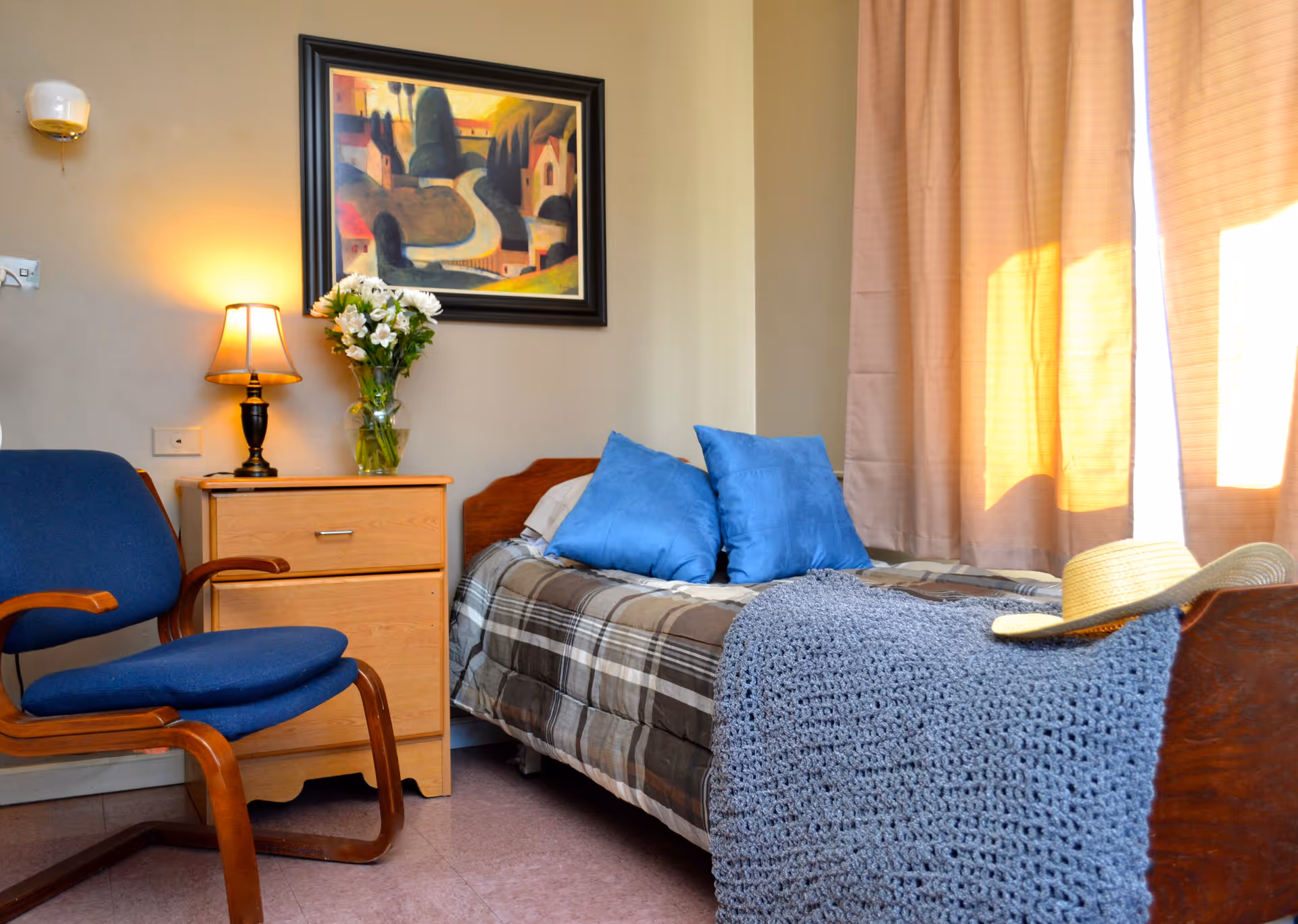 A cozy bedroom with a single bed covered in a plaid blanket and two blue pillows. A knitted gray throw and a straw hat rest on the bed. Next to the bed is a wooden nightstand with a lamp and a vase of white flowers. A blue cushioned chair with wooden armrests is positioned beside the nightstand. A framed painting hangs on the wall above the nightstand, and beige curtains cover the window, allowing warm sunlight to filter into the room.