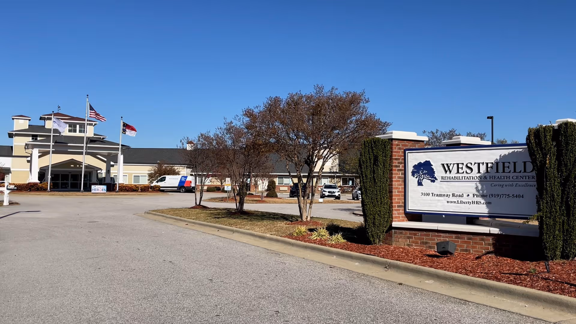 Exterior view of Westfield Rehabilitation & Health Center building with a clear blue sky. The entrance is visible with three flagpoles flying flags, and a parking area with some vehicles. A large sign with the facility name and contact information is in the foreground on the right side, surrounded by landscaping with small trees and bushes.