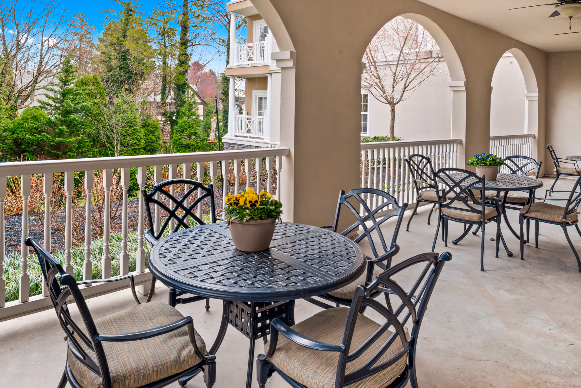 Covered outdoor patio area with multiple round metal tables and cushioned chairs. Each table has a small potted plant with flowers. The patio has arched openings and overlooks a landscaped garden with trees and shrubs.