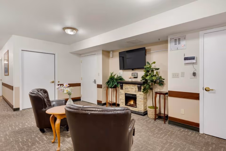 A cozy common area with two brown leather armchairs around a small wooden table with a vase of flowers. A wall-mounted flat-screen TV is above a stone fireplace with a small fire. There are two potted plants on either side of the fireplace, and three white doors are visible in the background. The room has beige walls with a wooden chair rail and carpeted floor.