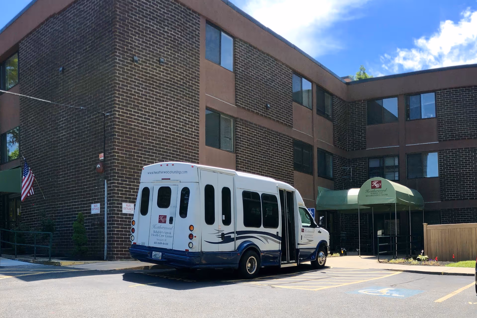 Exterior view of Heatherwood Rehabilitation & Health Care Center showing a brick building with multiple windows. A white and blue shuttle bus with the facility's name and contact information is parked in front near the main entrance, which has a green awning. An American flag is visible on the left side of the building.