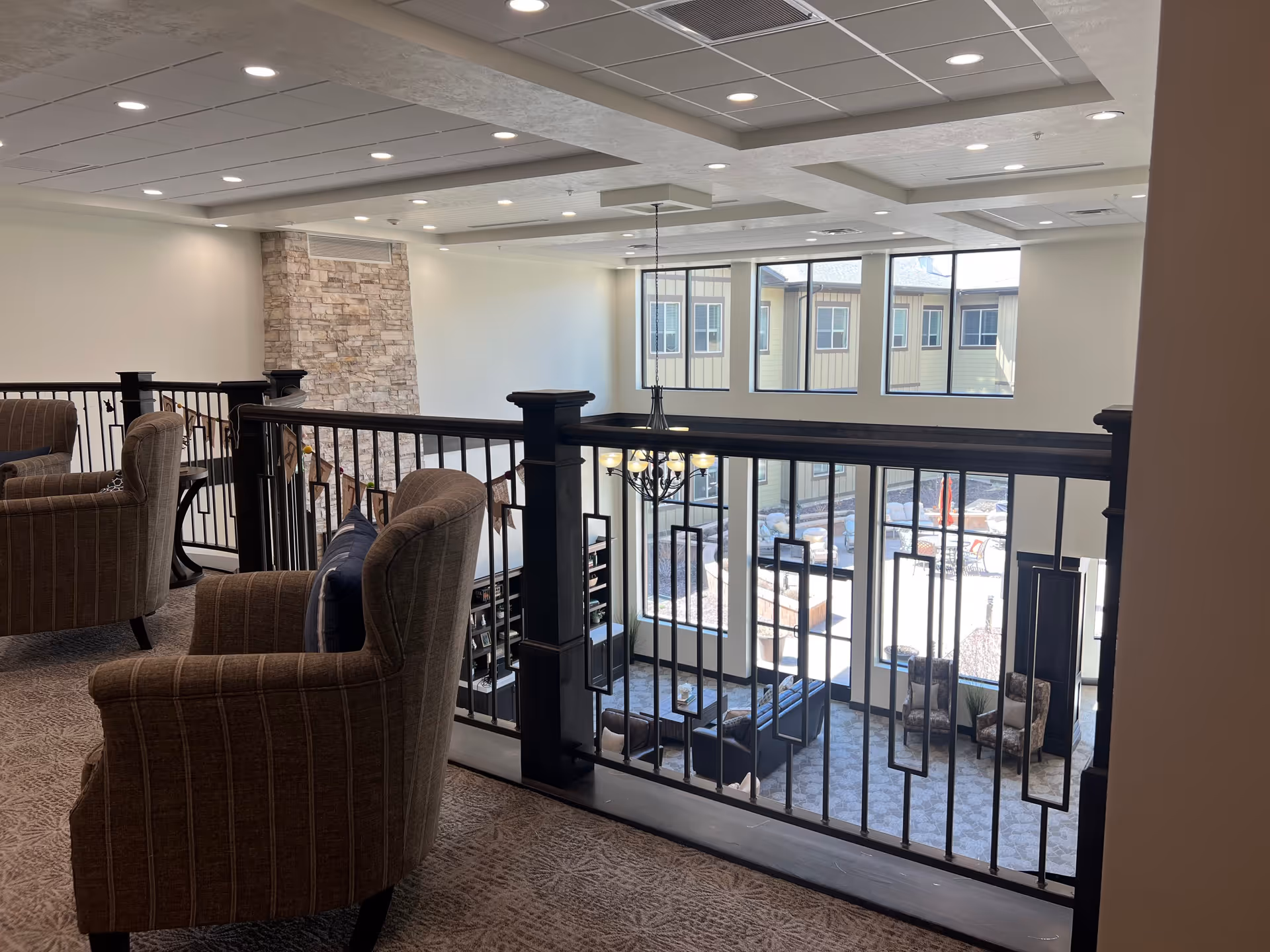 Interior view of a senior living facility lounge area with comfortable armchairs on a carpeted upper level overlooking a spacious common area below. The common area features large windows, a chandelier, and seating arrangements including sofas and armchairs. The design includes a stone accent wall and decorative railing.