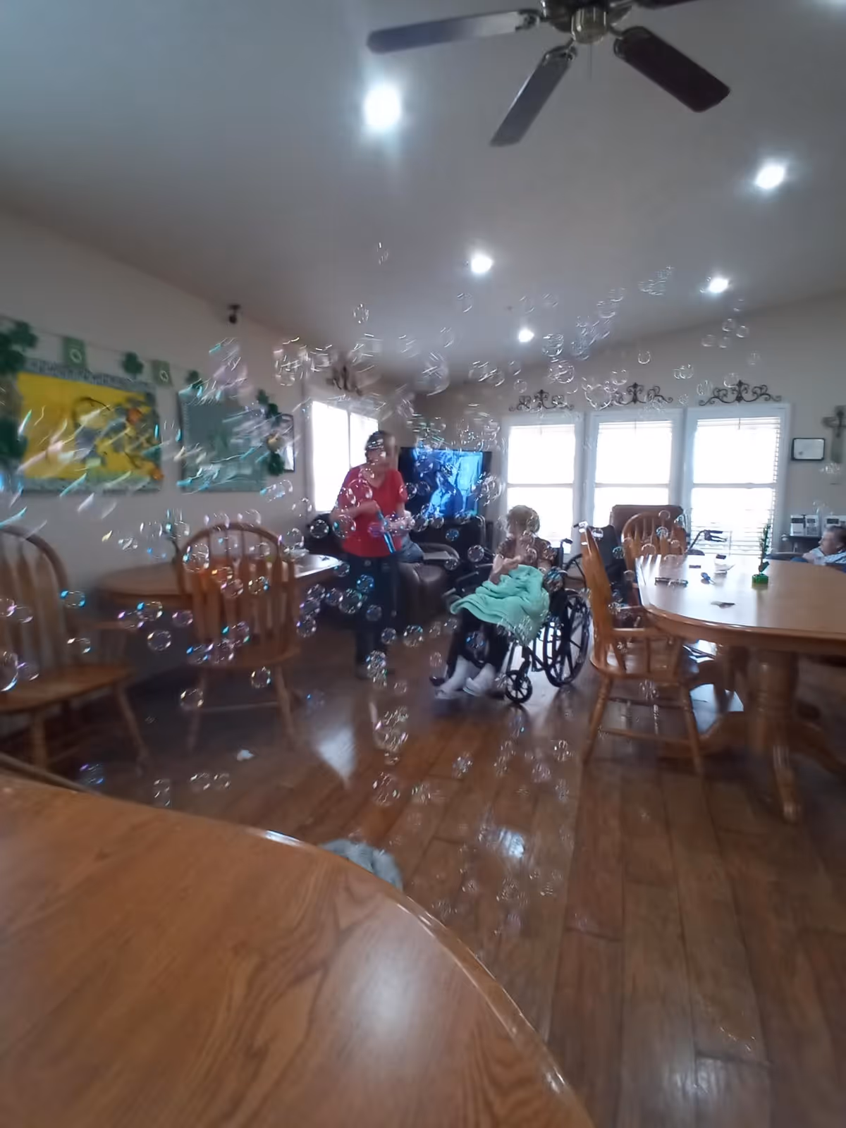 Communal dining/activity room with tables, chairs, a TV, a staff member and a resident in a wheelchair surrounded by many floating bubbles.