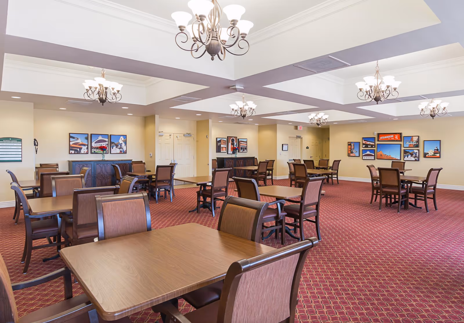 A spacious dining room with multiple wooden tables and chairs arranged neatly on a red patterned carpet. The room has cream-colored walls adorned with framed pictures and is illuminated by several elegant chandeliers hanging from a coffered ceiling.