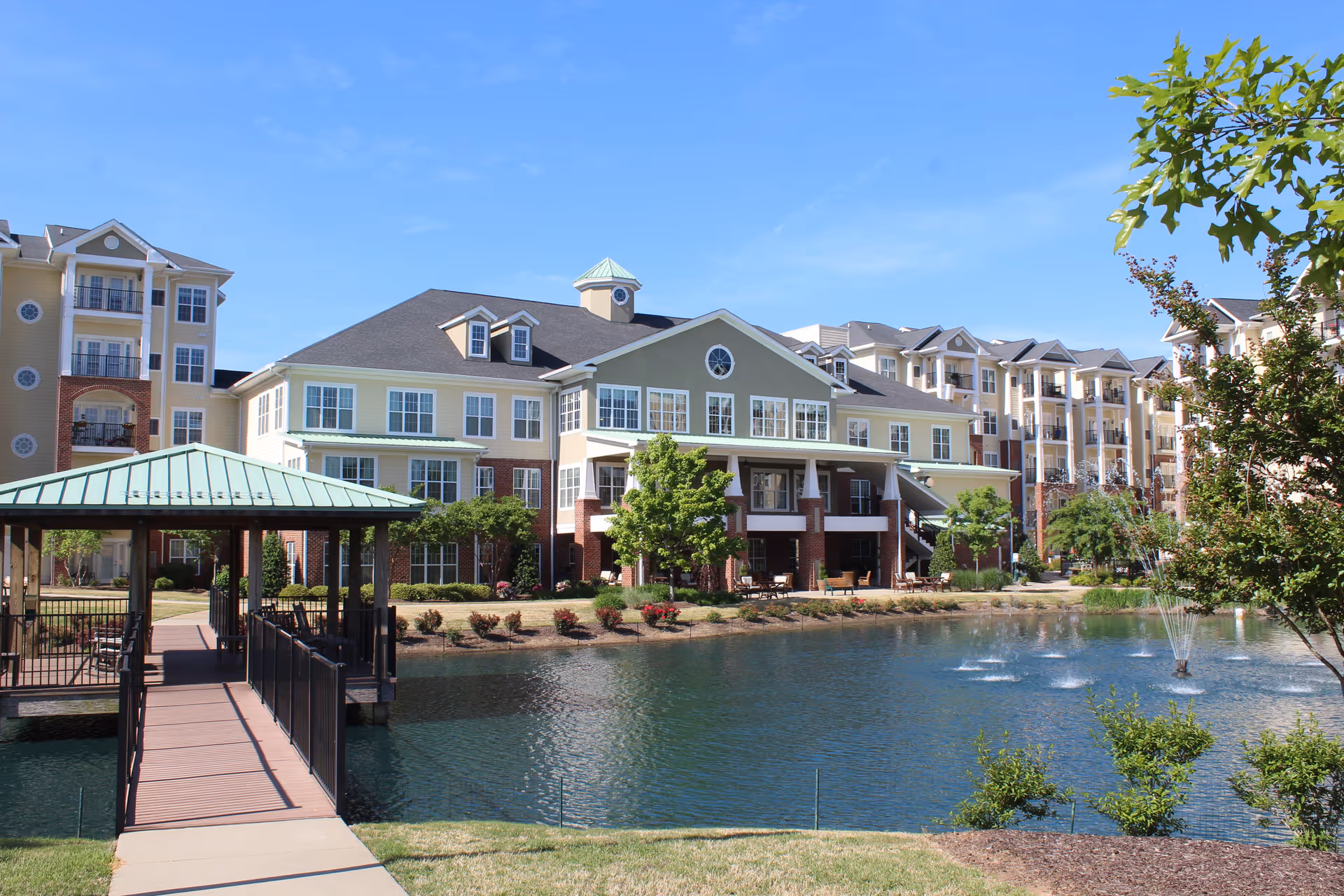 Exterior view of Cambridge Village of Apex senior living facility showing a large multi-story building with balconies, a pond with fountains, a covered wooden walkway leading over the water, and landscaped greenery under a clear blue sky.