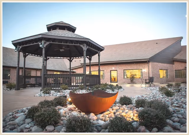 Outdoor courtyard area at Mira Vista Court featuring a large wooden gazebo with a ceiling fan, surrounded by landscaping with rocks and bushes. In the foreground, there is a rust-colored decorative bowl with lighting. The background shows a brick building with several windows reflecting the light.