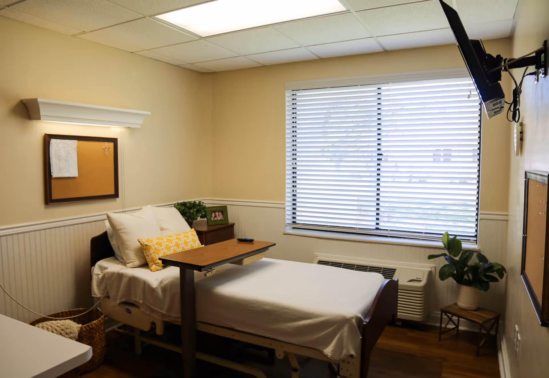 A small, cozy bedroom in a healthcare facility with a single hospital bed covered in white linens and pillows, including a yellow decorative pillow. A wooden overbed table is positioned over the bed. The room has beige walls with white wainscoting, a window with white blinds, a wall-mounted TV, a small wooden nightstand with a framed photo and a plant, and a basket on the floor.