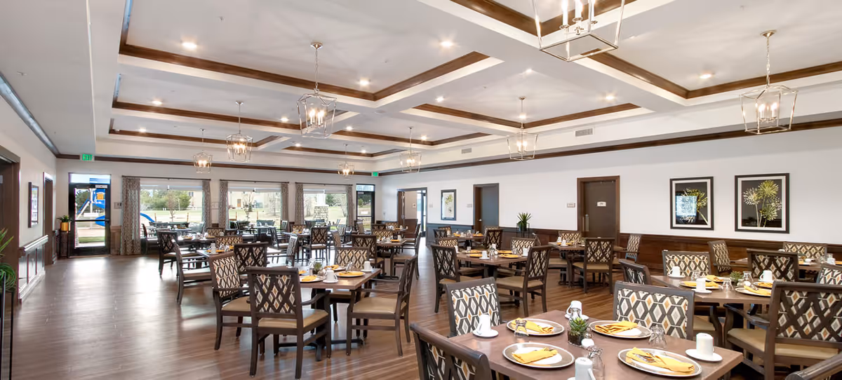 A spacious and well-lit dining room with multiple wooden tables and chairs arranged neatly. Each table is set with plates, cups, utensils, and yellow napkins. The room features a coffered ceiling with modern pendant lights and large windows that provide a view of an outdoor playground area. The walls are decorated with framed botanical artwork and there are several doors leading to other areas.